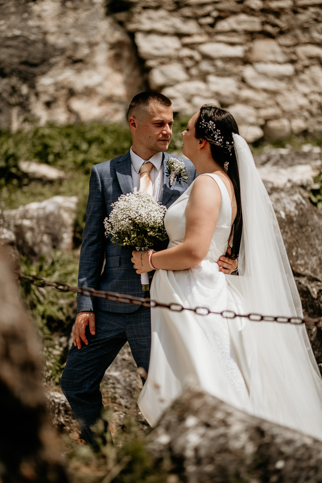 Svadobná fotografia Dominiky a Mariána pred impozantnými hradbami Spišského hradu, so širokým úsmevom a láskyplným pohľadom.